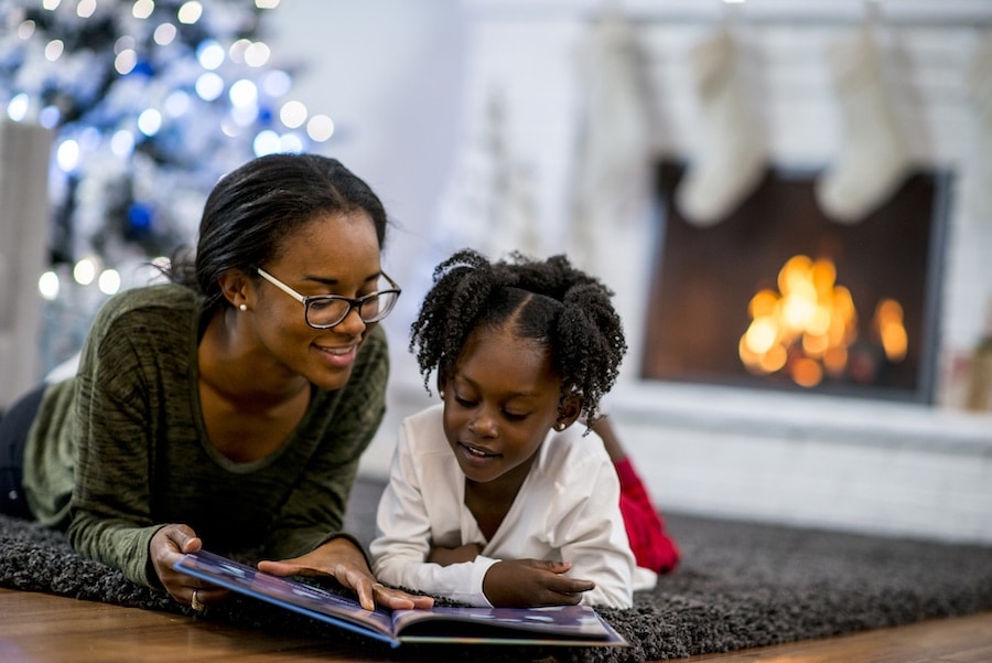 A mother and daughter of African descent are in indoors in their living room during Christmas time. They are wearing warm