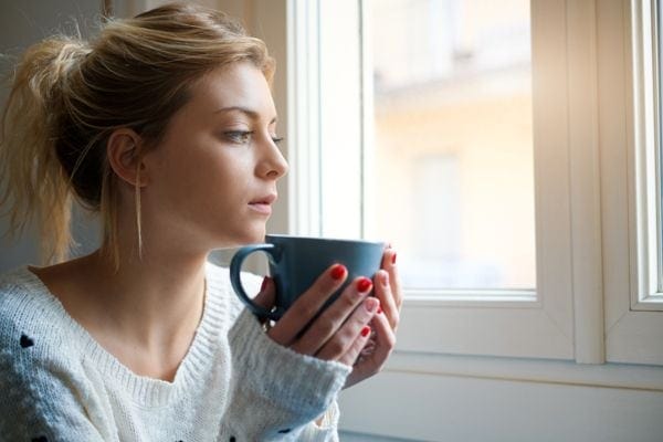 A woman looks out a window while holding a cup. Should I Get an 80% or 95% Furnace?